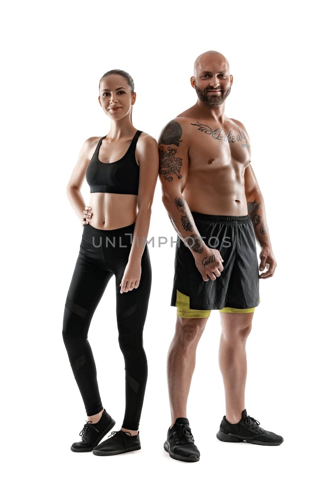 Athletic bald, tattooed male in black shorts and sneakers with beautiful brunette female in leggings and top are posing isolated on white background and looking at the camera. Fitness couple, chic muscular bodies, gym concept. The love story.