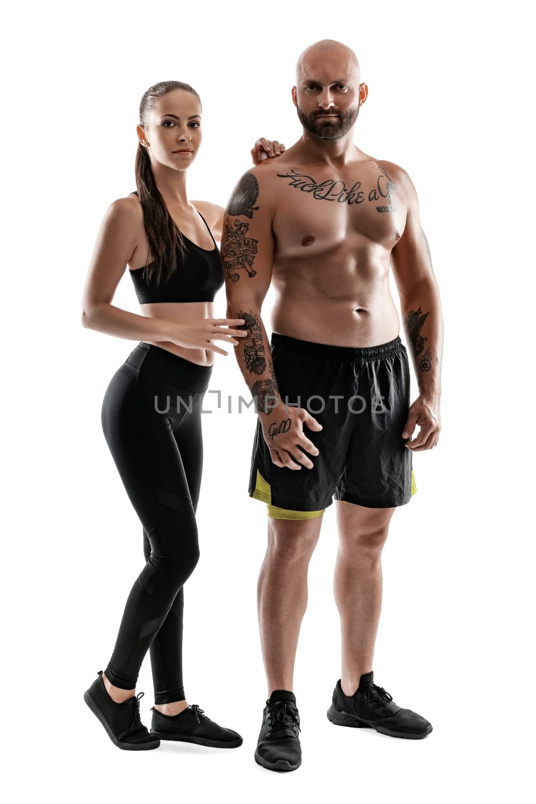 Athletic bald, tattooed fellow in black shorts and sneakers with beautiful brunette lady in leggings and top are posing isolated on white background and looking at the camera. Fitness couple, chic muscular bodies, gym concept. The love story.