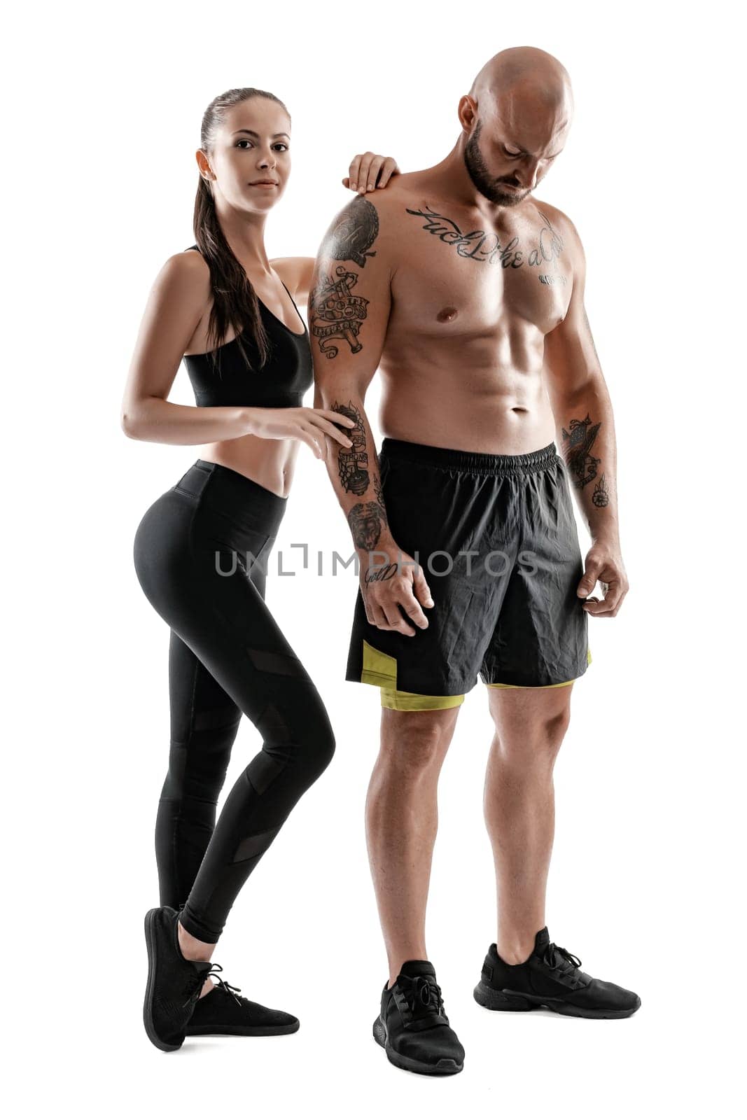 Strong bald, tattooed male in black shorts and sneakers with charming brunette female in leggings and top are posing isolated on white background and looking at the camera. Fitness couple, chic muscular bodies, gym concept. The love story.