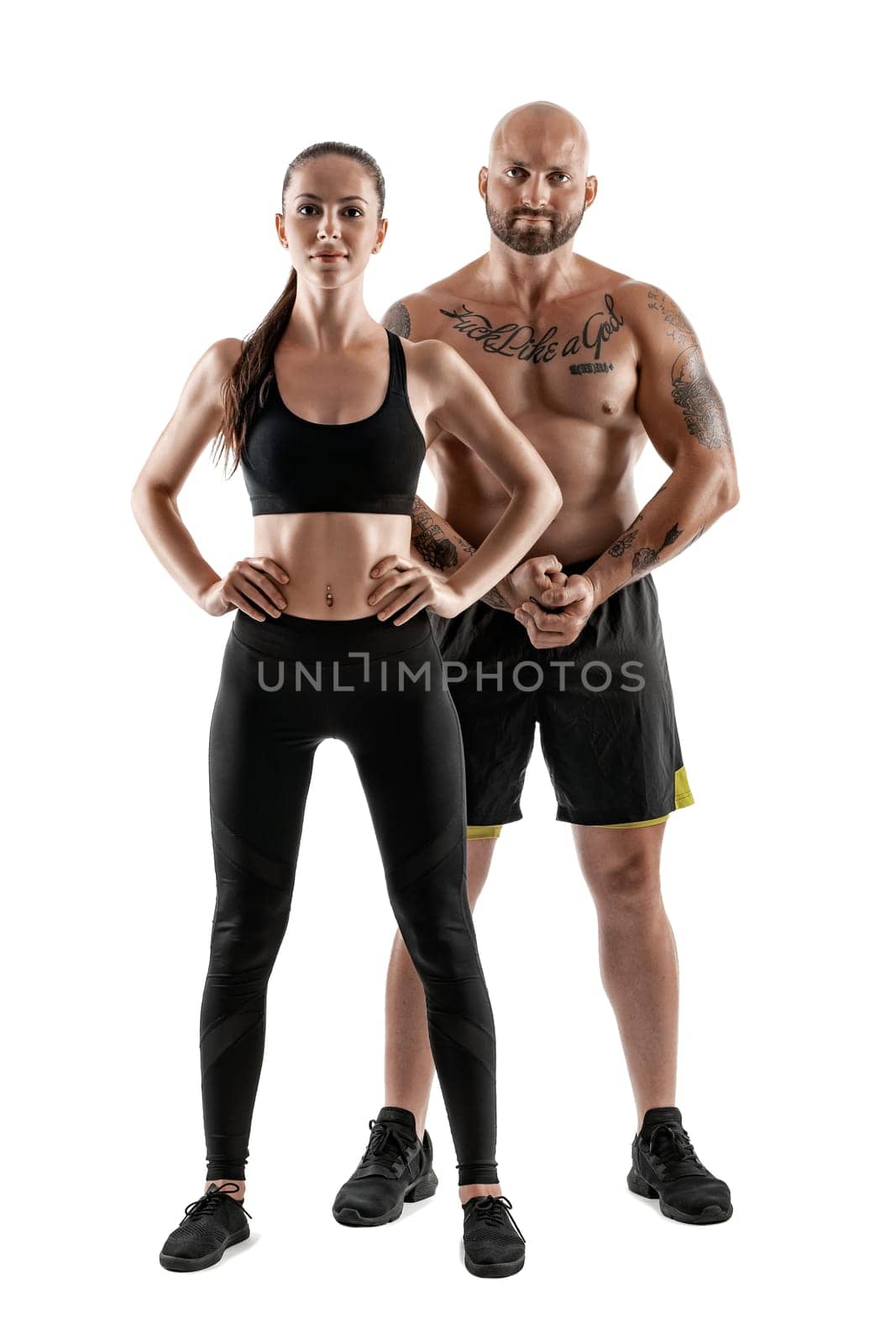Athletic bald, tattooed male in black shorts and sneakers with gorgeous brunette female in leggings and top are posing isolated on white background and looking at the camera. Fitness couple, chic muscular bodies, gym concept. The love story.