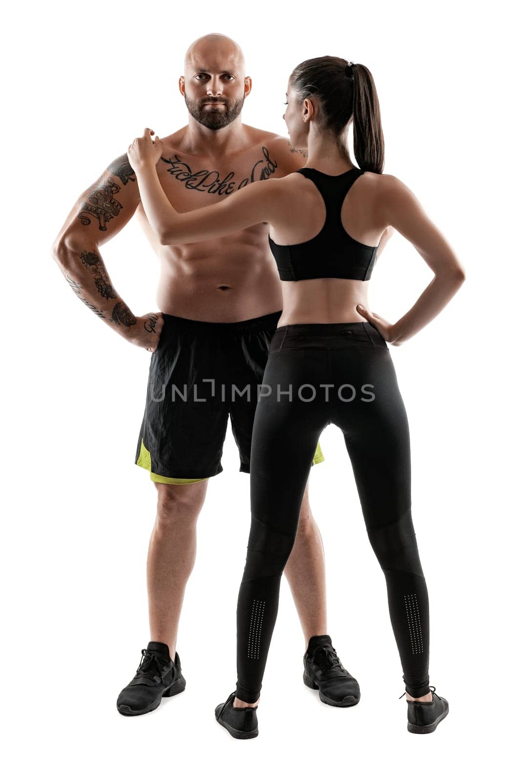 Handsome bald, tattooed male in black shorts and sneakers with attractive brunette female in leggings and top, standing back,are posing isolated on white background and looking at the camera. Fitness couple, chic muscular bodies, gym concept. The love story.