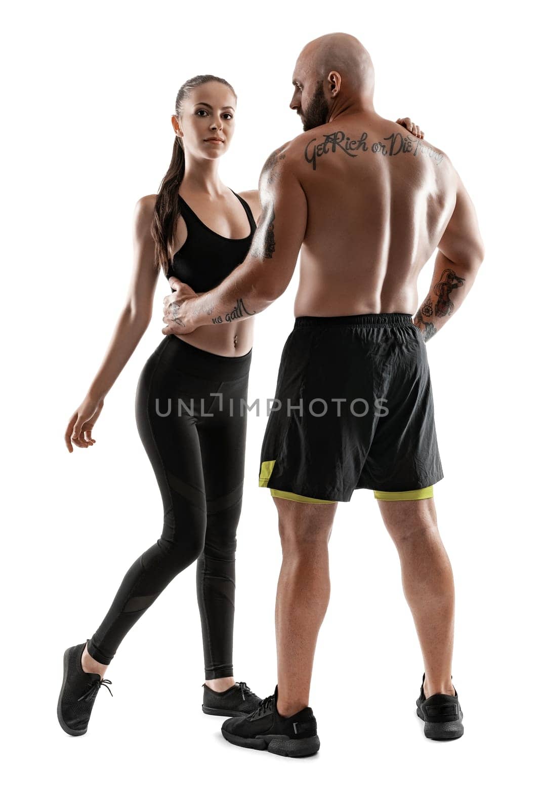 Good-looking bald, tattooed man in black shorts and sneakers, standing back, with alluring brunette maiden in leggings and top are posing isolated on white background and looking at the camera. Fitness couple, chic muscular bodies, gym concept. The love story.