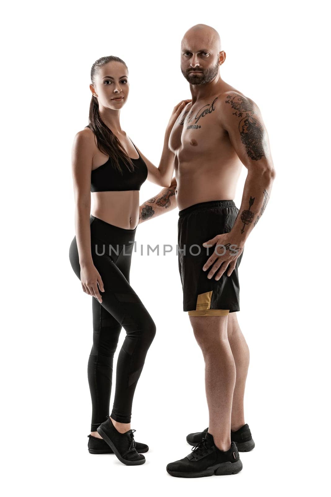 Stately bald, tattooed male in black shorts and sneakers with pretty brunette female in leggings and top are posing standing sideways isolated on white background and looking at the camera. Fitness couple, chic muscular bodies, gym concept. The love story.