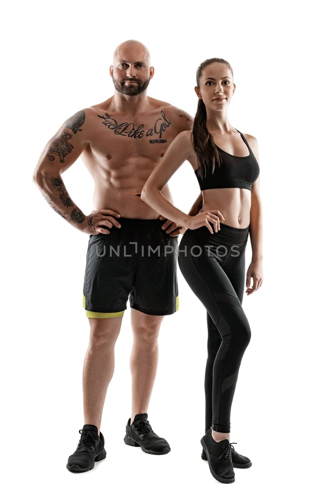 Athletic bald, tattooed man in black shorts and sneakers with cute brunette woman in leggings and top are posing isolated on white background and looking at the camera. Fitness couple, chic muscular bodies, gym concept. The love story.