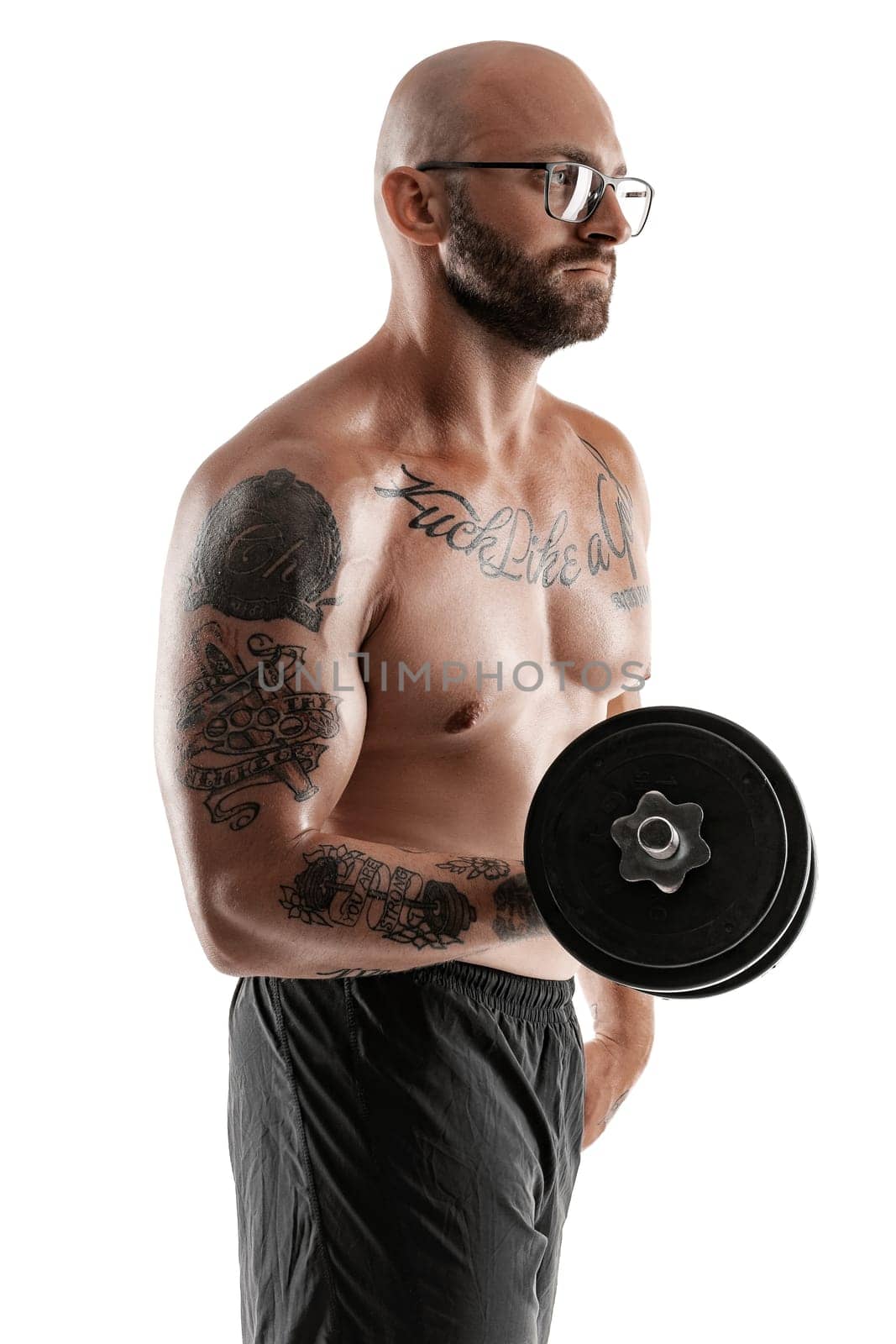 Handsome bald, bearded, tattooed fellow in glasses, black shorts is posing holding a dumbbell in his hand isolated on white background and looking away. Chic muscular body, fitness, gym, healthy lifestyle concept. Close-up portrait.