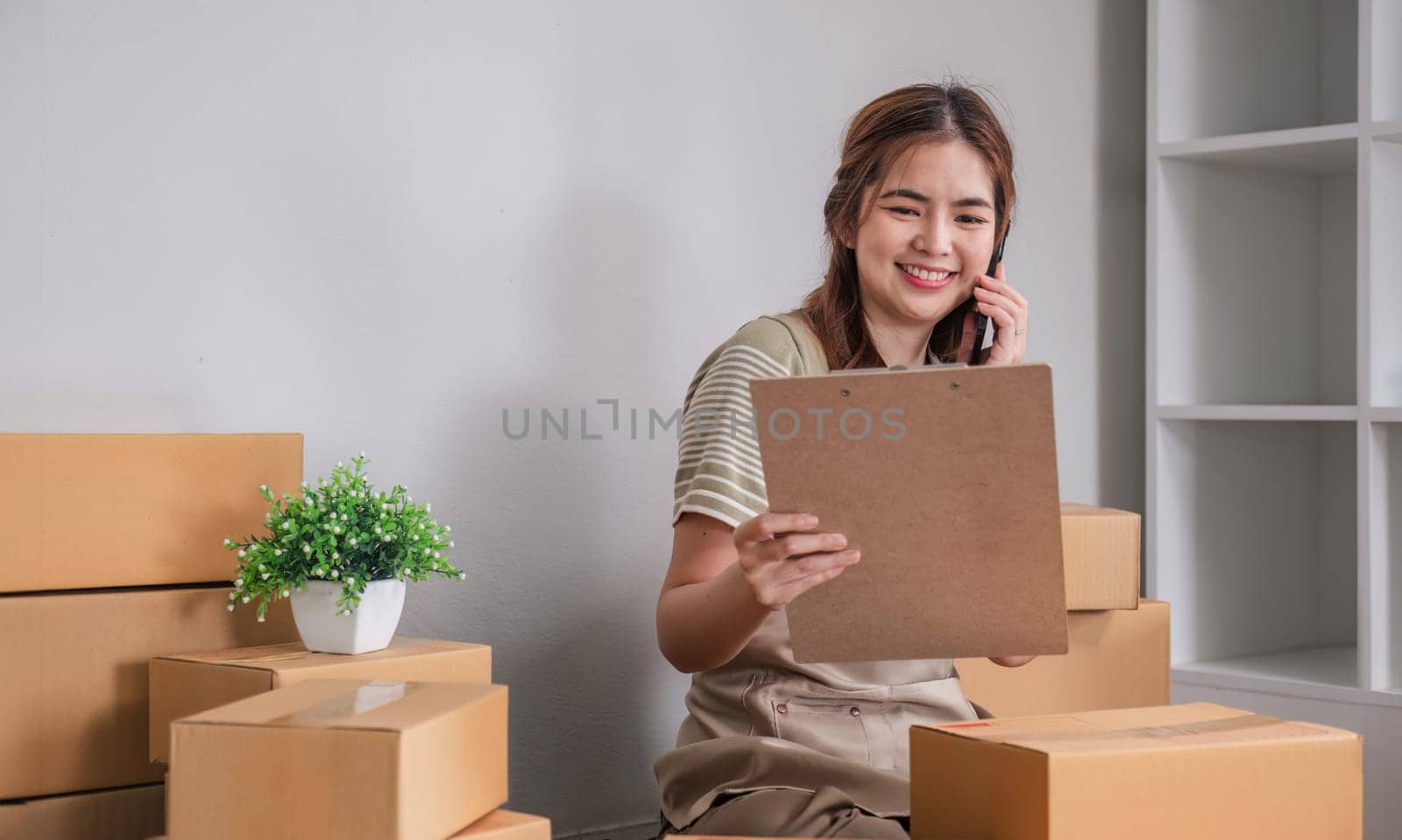 Small business entrepreneur SME freelance woman using phone call receive from customer checking product on stock at home office, online marketing packaging delivery box, SME e-commerce concept. by wichayada