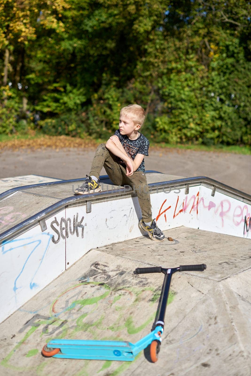 Boy on scooter makes a trick and enjoying his riding in the skate park at cloudy spring day. Young man doing trick on the kick scooter in the park. Full body little boy on the kick scooter on sunny autumn day in skate park. From below of active teen boy with kick scooter standing on ramp in skate park while preparing for performing trick in sunny spring day.