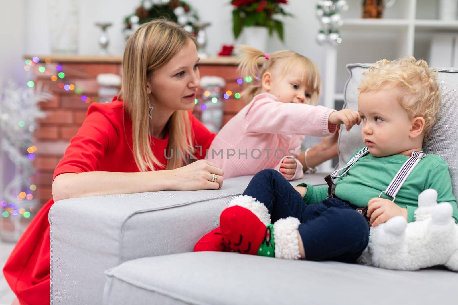 Mother, daughter and son against the background of a brick fireplace. The boy sits on the couch. Next to the couch crouches a woman. Next to the woman stands a little girl and extends her hand to her brother. Blurred background.