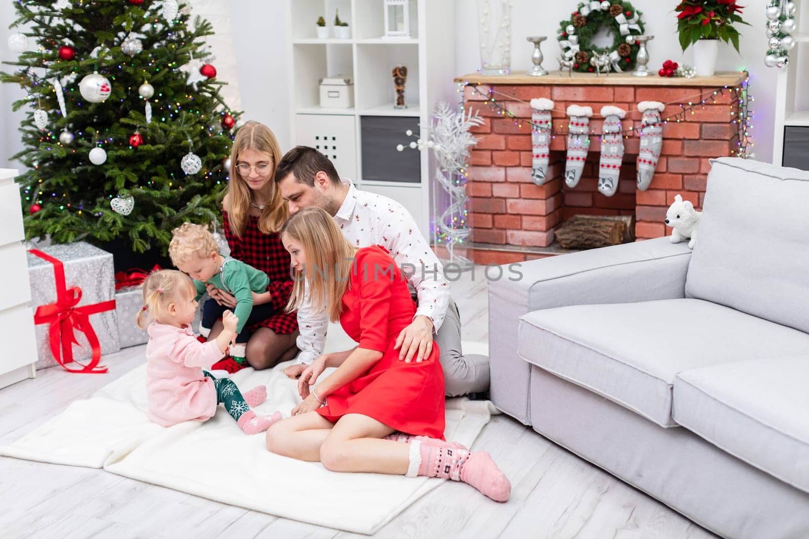 Two women, a man and two children spend Christmas together. The family sits on a white carpet in front of the fireplace. The fireplace is decorated with colorful lights. Presents lie under the large Christmas tree.