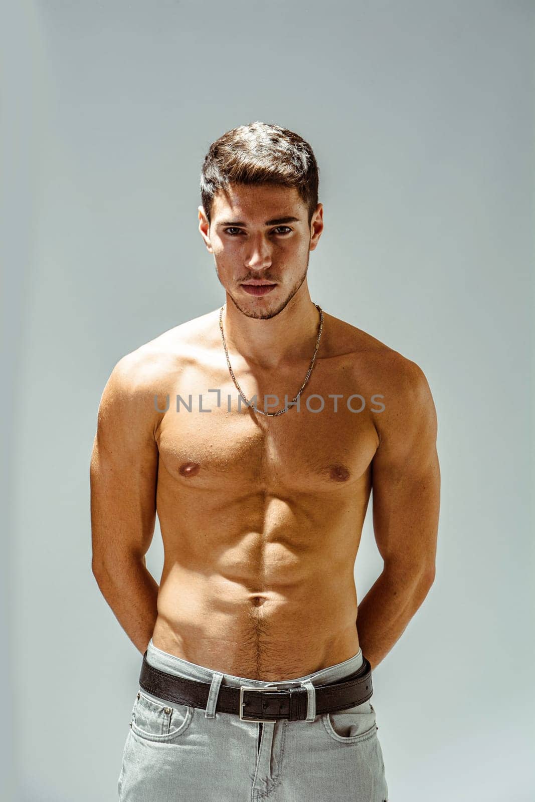 A shirtless handsome young man posing for a picture showing muscular torso, looking at camera, wearing grey jeans