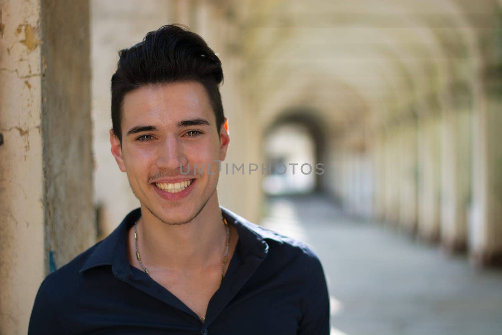 A young man is smiling for the camera. Photo of a young man with a bright smile, outdoor under a colonnade in European city. A cheerful young adult smiling with eye contact.