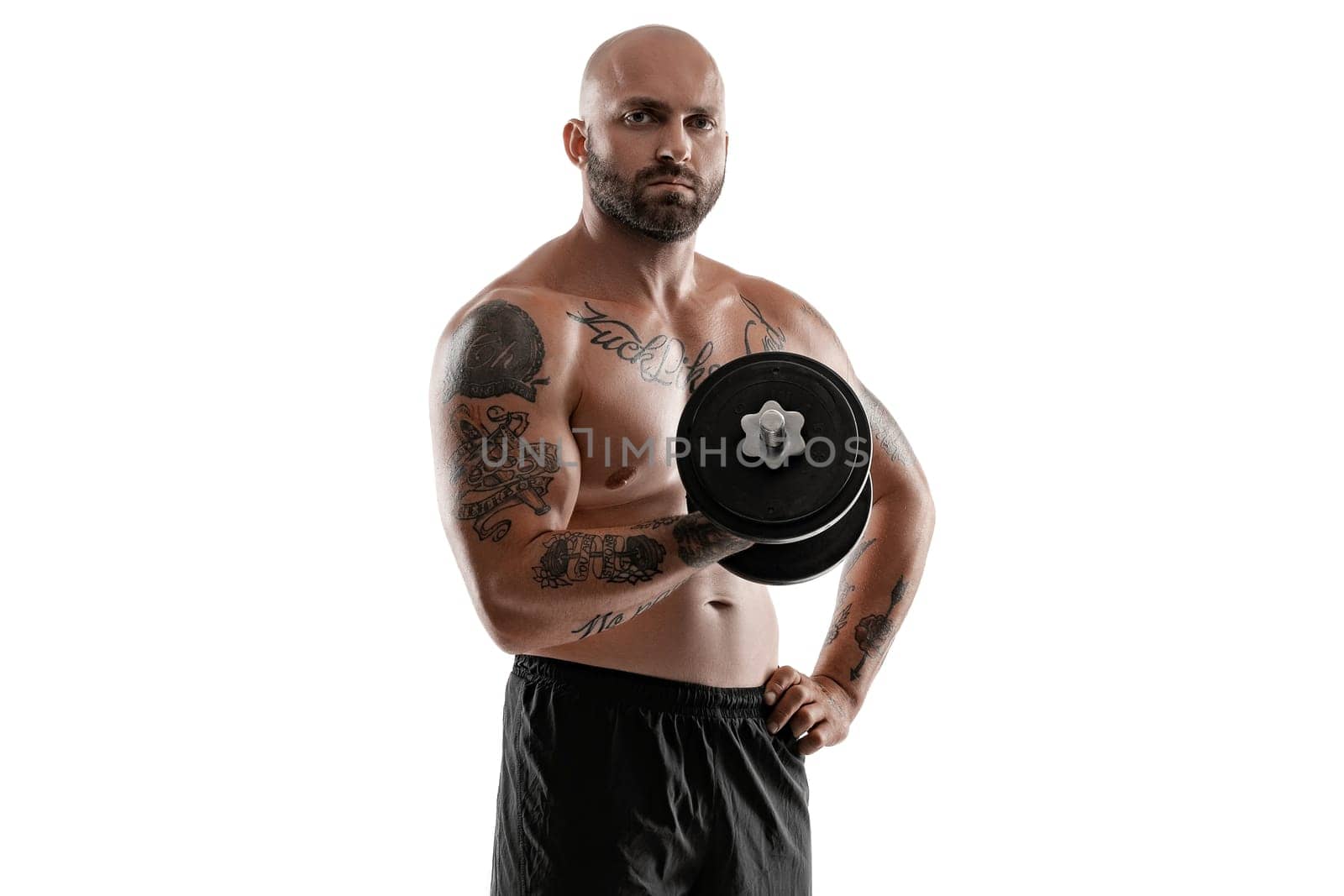 Good-looking bald, bearded, tattooed male in black shorts is posing holding a dumbbell in his hand and looking at the camera, isolated on white background . Chic muscular body, fitness, gym, healthy lifestyle concept. Close-up portrait.