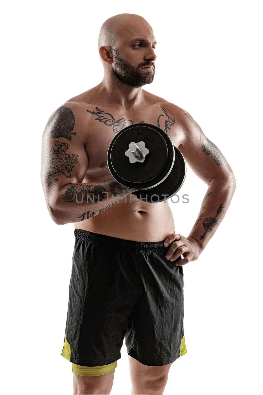 Good-looking bald, bearded, tattooed man in black shorts is posing holding a dumbbell in his hand and looking away, isolated on white background . Chic muscular body, fitness, gym, healthy lifestyle concept. Close-up portrait.