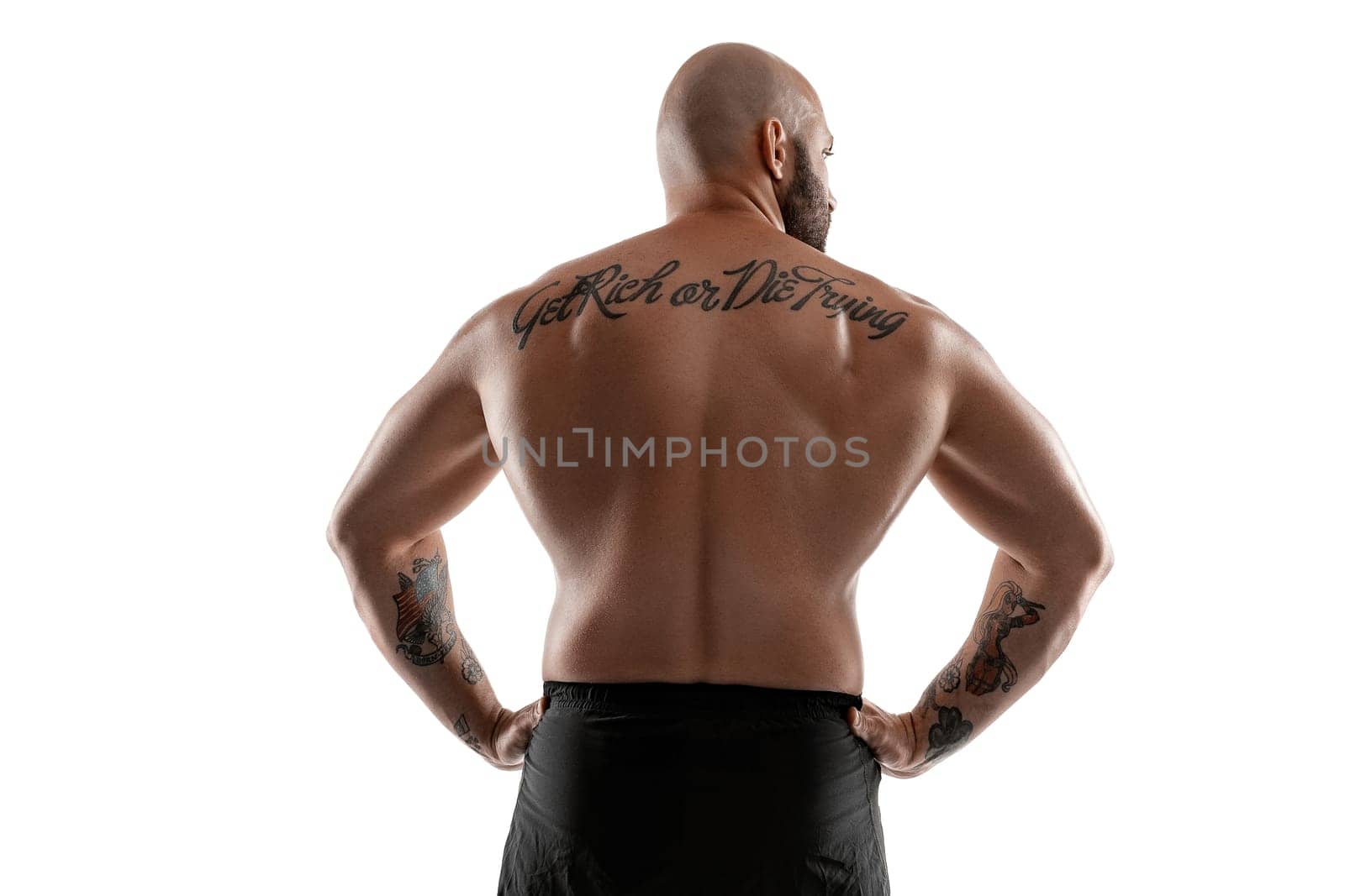 Stately bald, bearded, tattooed man in black shorts is posing standing back to the camera and showing his muscles, isolated on white background. Chic muscular body, fitness, gym, healthy lifestyle concept. Close-up portrait.