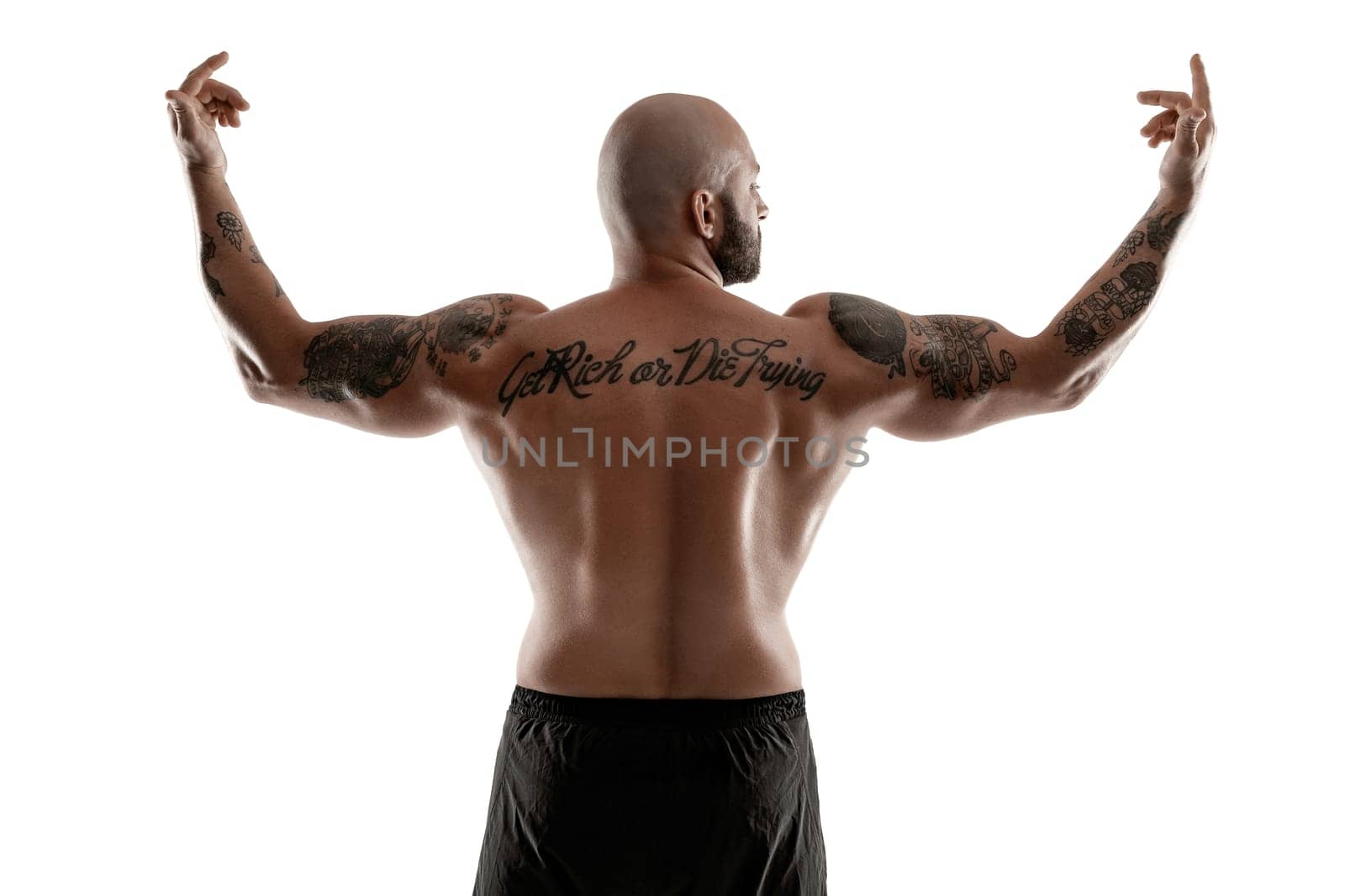 Stately bald, bearded, tattooed guy in black shorts is posing standing back to the camera, raised his hands and showing muscles, isolated on white background. Chic muscular body, fitness, gym, healthy lifestyle concept. Close-up portrait.