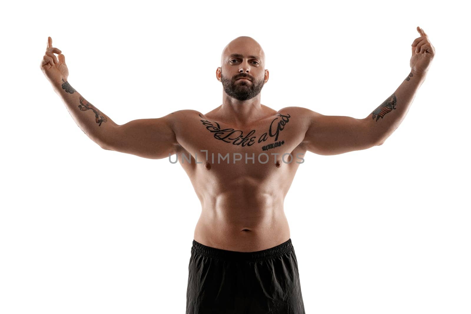 Stately bald, bearded, tattooed fellow in black shorts is posing isolated on white background, raised his hands and showing muscles, looking at the camera. Chic muscular body, fitness, gym, healthy lifestyle concept. Close-up portrait.
