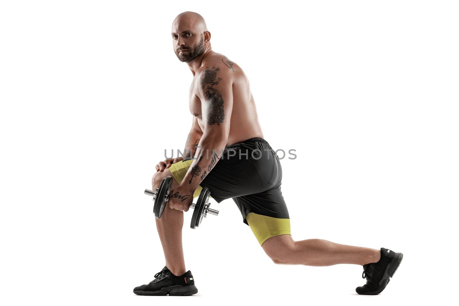 Strong bald, bearded, tattooed male in black shorts and sneakers is posing with a dumbbell in his hand, standing sideways and looking at the camera, isolated on white background. Chic muscular body, fitness, gym, healthy lifestyle concept. Full length portrait.