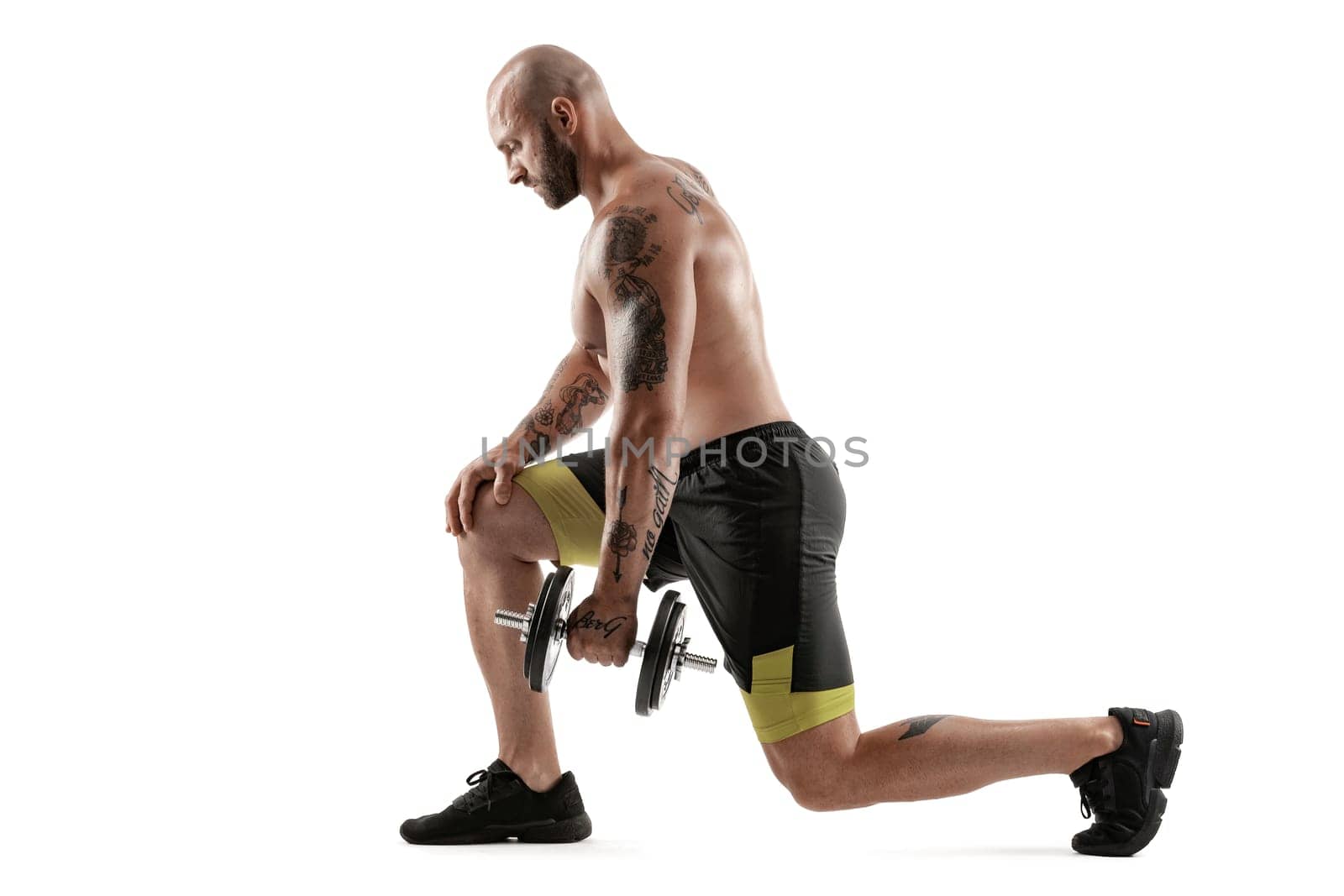 Strong bald, bearded, tattooed fellow in black shorts and sneakers is posing with a dumbbell in his hand, standing sideways isolated on white background. Chic muscular body, fitness, gym, healthy lifestyle concept. Full length portrait.