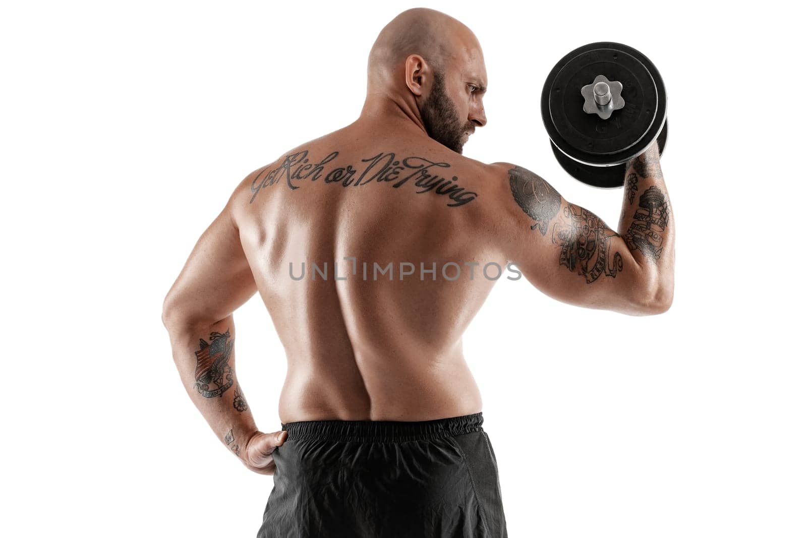 Masculine bald, bearded, tattooed man in black shorts is posing with a dumbbell in his hand, standing back, isolated on white background. Chic muscular body, fitness, gym, healthy lifestyle concept. Close-up portrait.