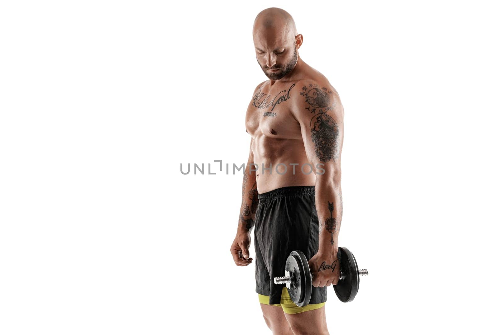 Masculine bald, bearded, tattooed fellow in black shorts is posing standing sideways, with a dumbbell in his hand, isolated on white background. Chic muscular body, fitness, gym, healthy lifestyle concept. Close-up portrait.