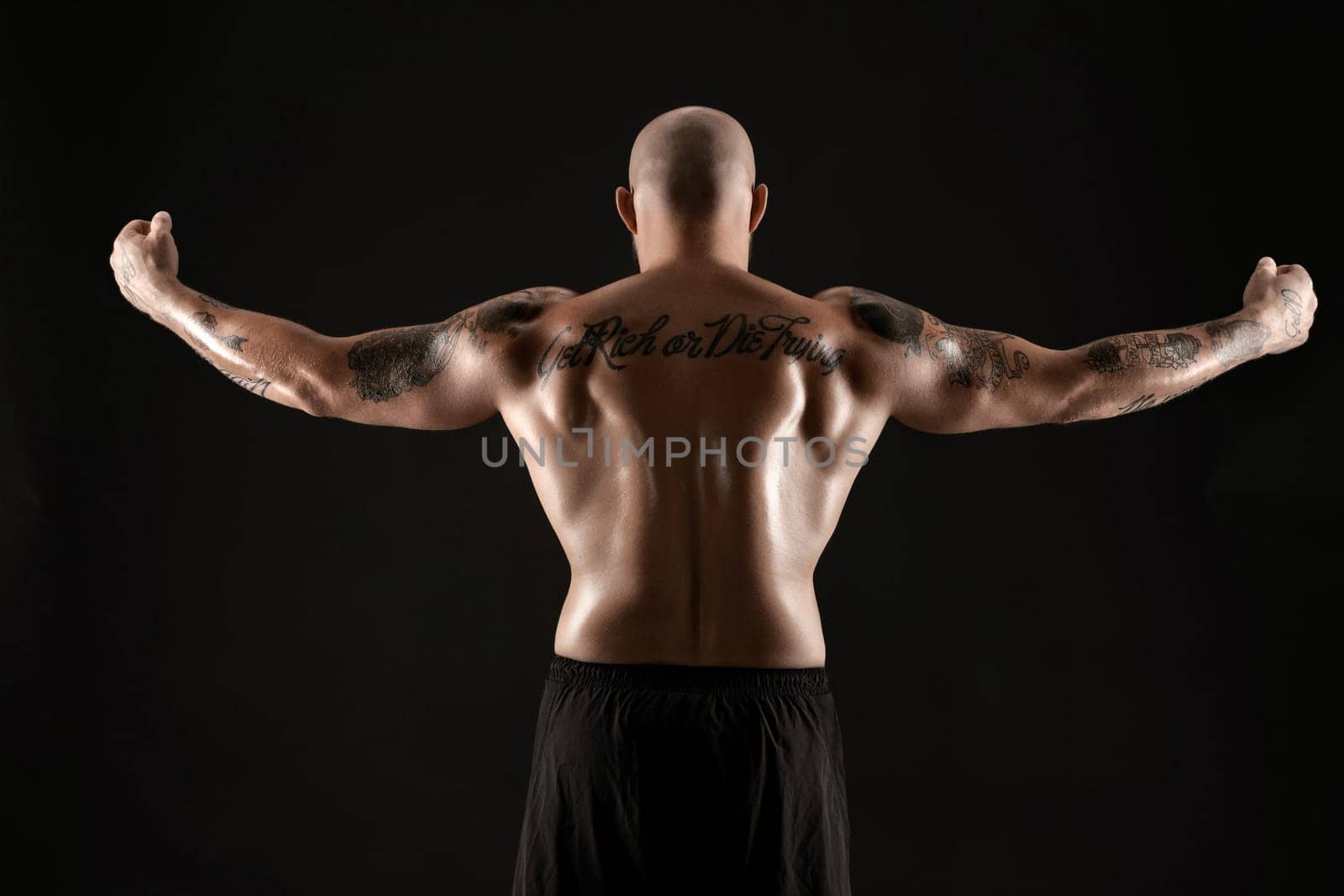 Athletic bald, bearded, tattooed man in black shorts is posing standing back to the camera and showing his muscles, standing against a black background. Chic muscular body, fitness, gym, healthy lifestyle concept. Close-up portrait.