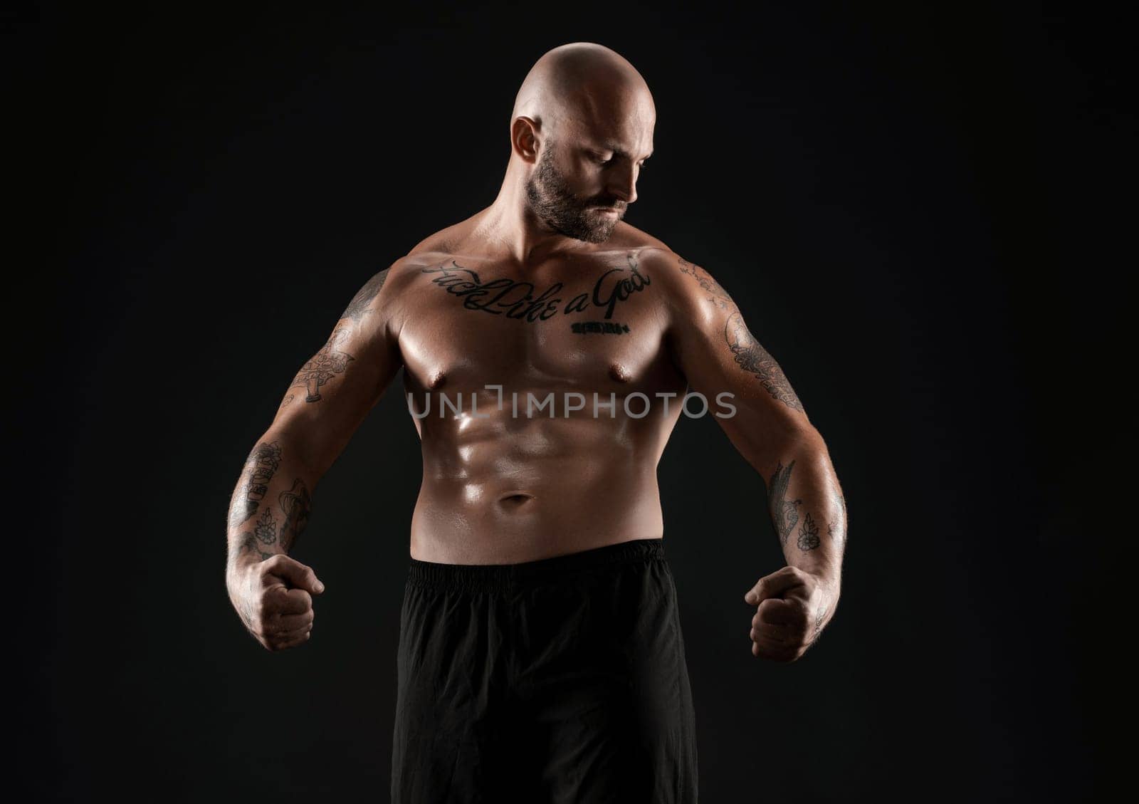 Athletic bald, bearded, tattooed person in black shorts is demonstating his muscles posing against a black background and looking down. Chic muscular body, fitness, gym, healthy lifestyle concept. Close-up portrait.