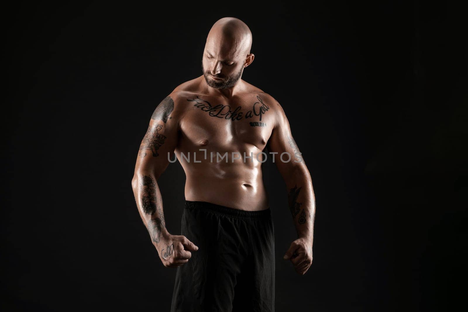 Handsome bald, bearded, tattooed man in black shorts is demonstating his muscles and looking at it while posing against a black background. Chic muscular body, fitness, gym, healthy lifestyle concept. Close-up portrait.