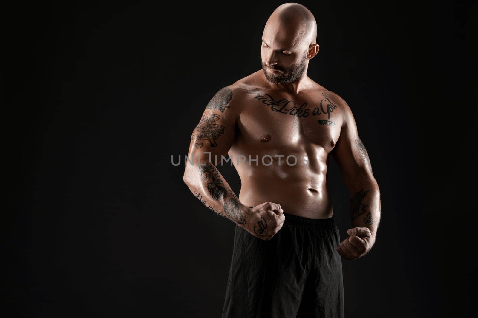 Athletic bald, bearded, tattooed man in black shorts is posing against a black background. Close-up portrait. by nazarovsergey