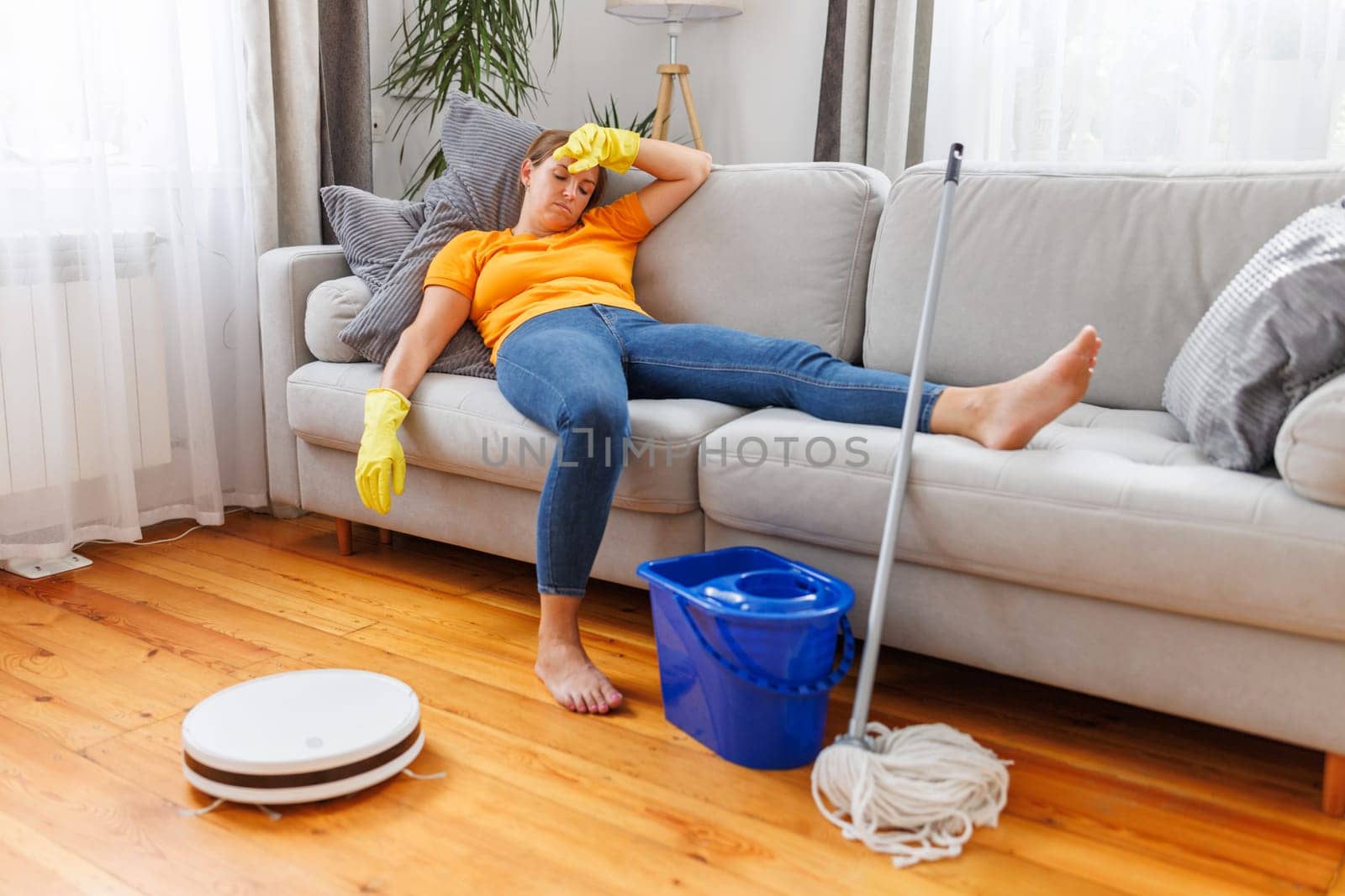 Robotic vacuum cleaner cleaning a room while tired woman relaxing on the sofa after doing housework. Concept of the advantages of modern cleaning technologies, buying a robot vacuum cleaner