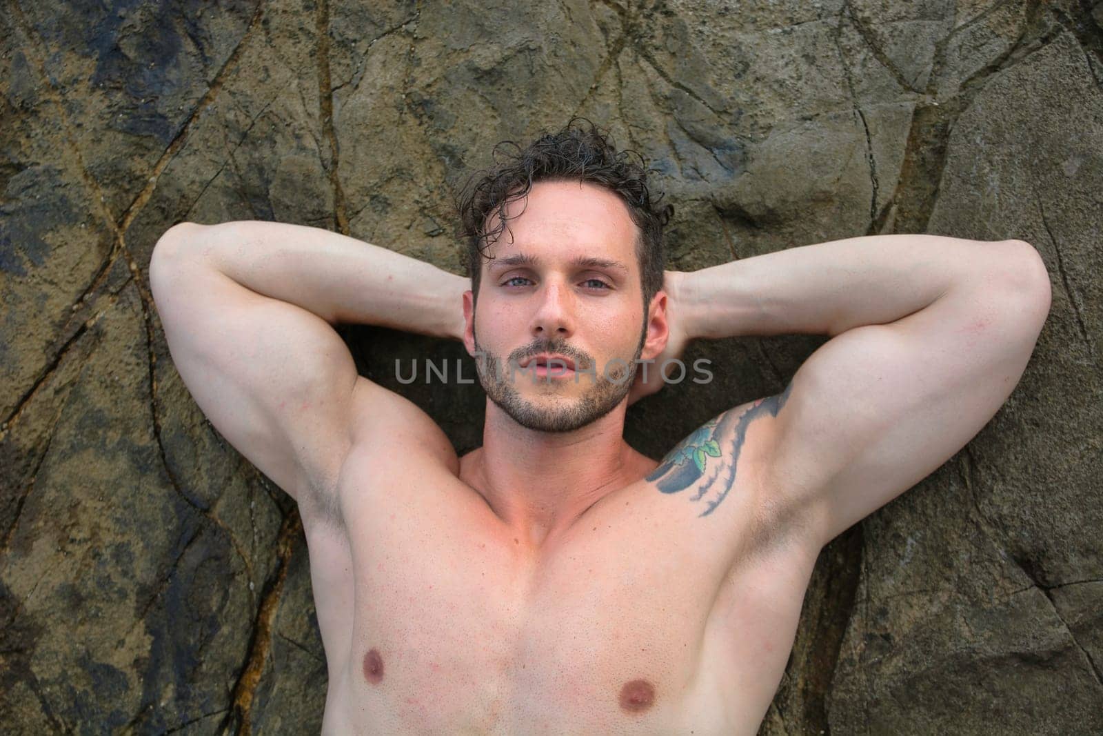 A shirtless muscular man laying on a rock next to the sea. Photo of a man relaxing by the water's edge on a sunny day