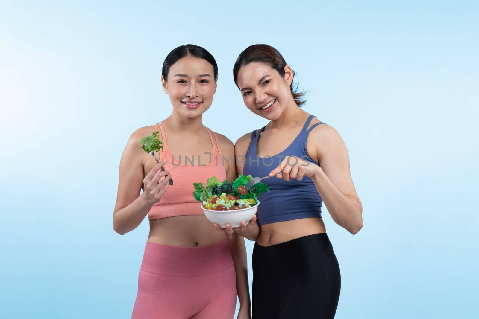 Two young sporty Asian women in sportswear holding salad bowl fill with fruit and vegetable. Natural youthful and fit body lifestyle people with balance nutrition on isolated background. Vigorous