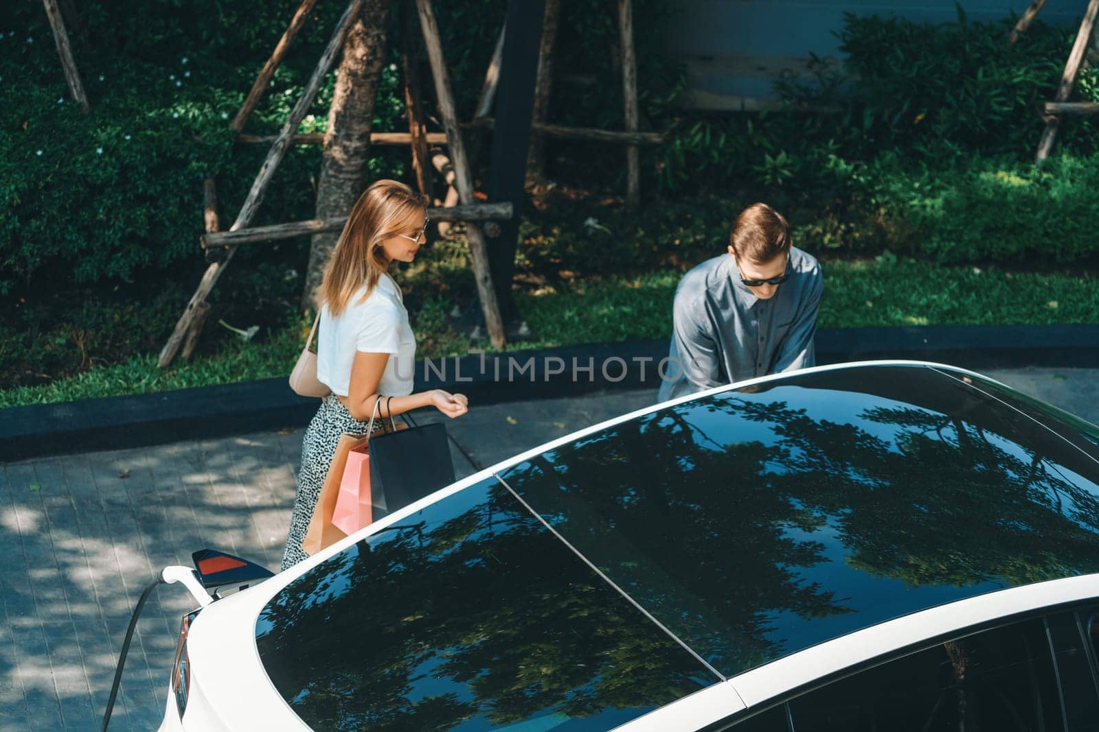Young couple travel with EV electric car charging in green sustainable city outdoor garden in summer shows urban sustainability lifestyle by green clean rechargeable energy of electric vehicle innards