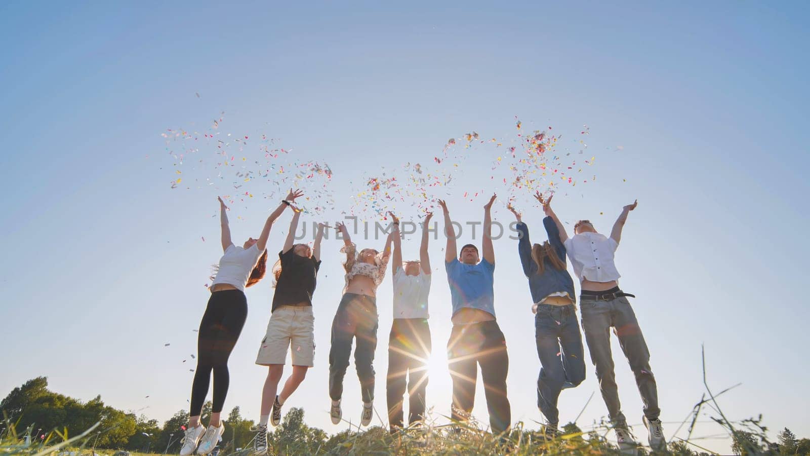 Friends toss colorful paper confetti from their hands against the rays of the evening sun