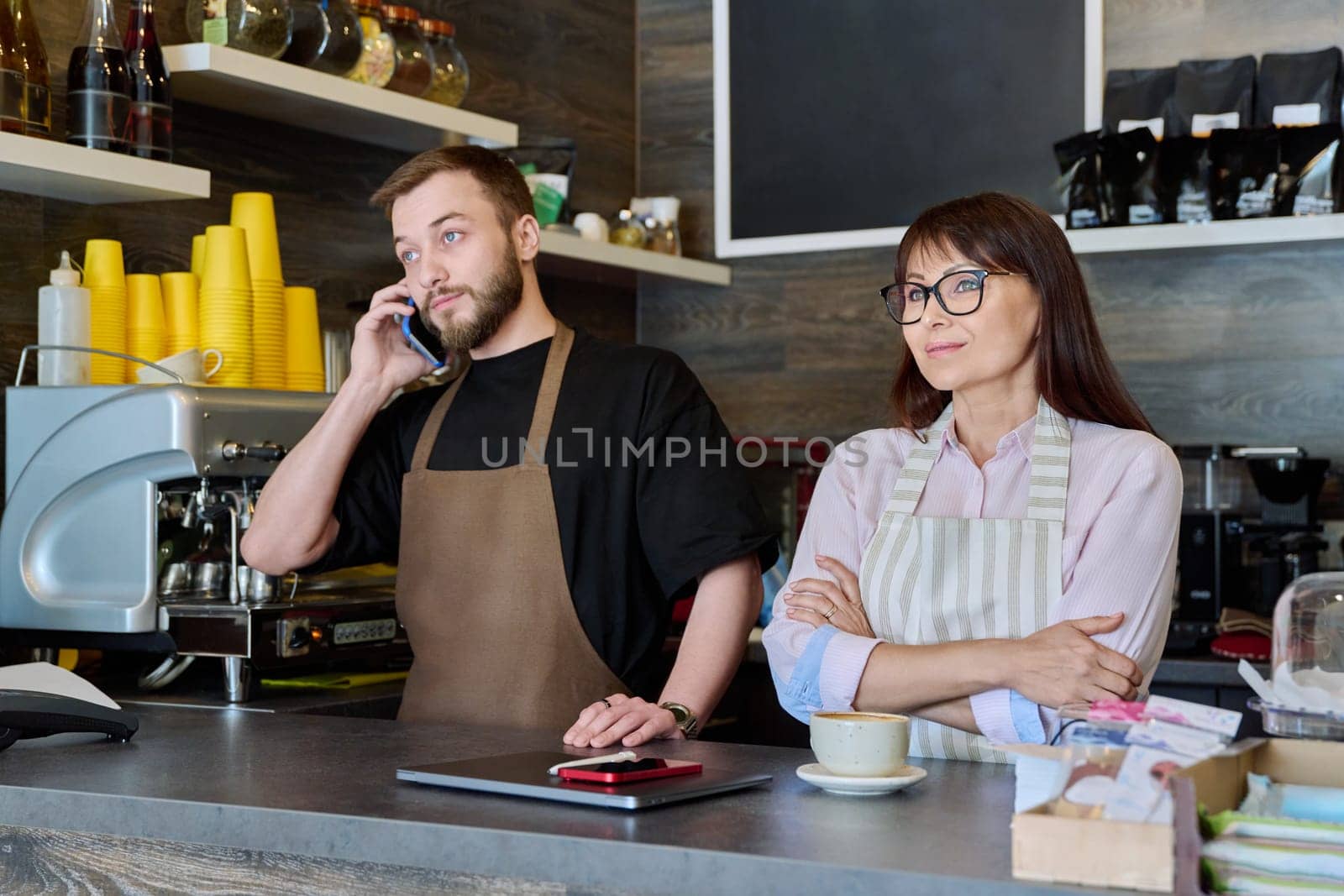 Team, partners, young male and mature woman talking working using phone standing behind bar in coffee shop. Team, small business, work, staff, cafe cafeteria restaurant, entrepreneurship concept