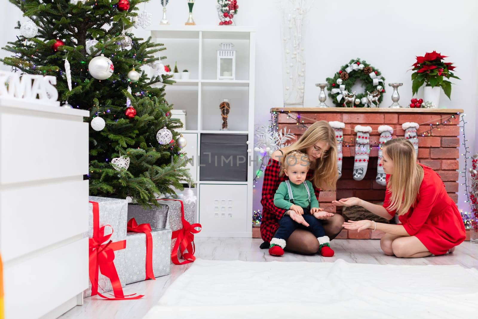 Two women talk by the fireplace. The women are sitting by a decorated Christmas tree. One of the women holds a little boy on her lap. One of the women is wearing glasses.