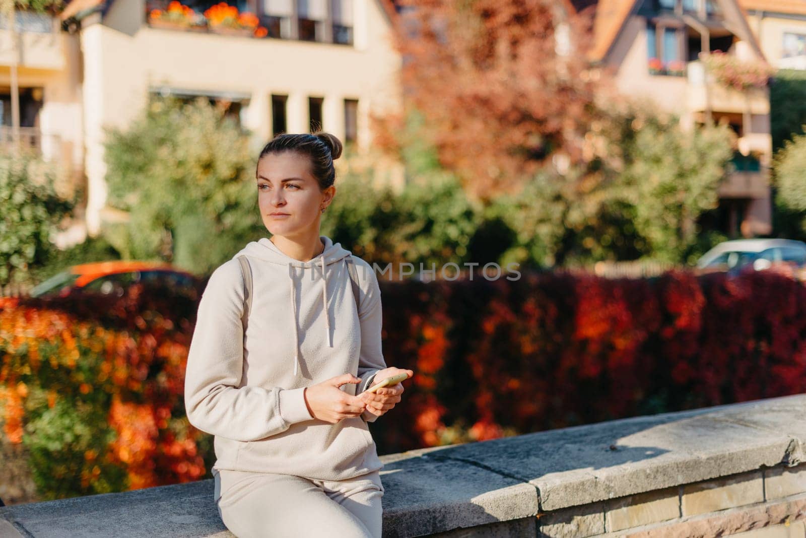 Young Fashionable Teenage Girl With Smartphone In Europian Park In Autumn Sitting At Smiling. Trendy Young Woman In Fall In Park Texting. Retouched, Vibrant Colors. Beautiful Blonde Teenage Girl Wearing Casual Modern Autumn Outfit Sitting In Park In Autumn. Retouched, Vibrant Colors, Brownish Tones. by Andrii_Ko