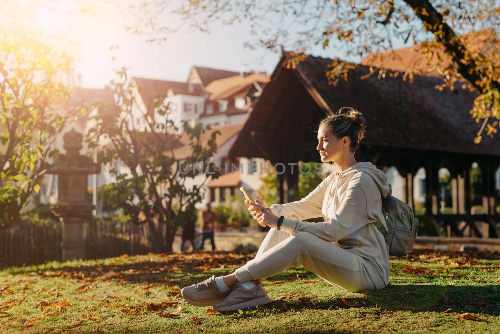 Young fashionable teenage girl with smartphone in park in autumn sitting at smiling. Trendy young woman in fall in park texting. Retouched, vibrant colors. Beautiful blonde teenage girl wearing casual modern autumn outfit sitting in park in autumn. Retouched, vibrant colors, brownish tones.