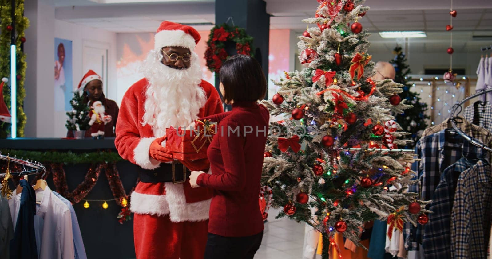 African american employee acting as Santa Claus sitting down next to Christmas tree with asian woman in xmas ornate clothing store. Fashion shop worker offering gift to customer