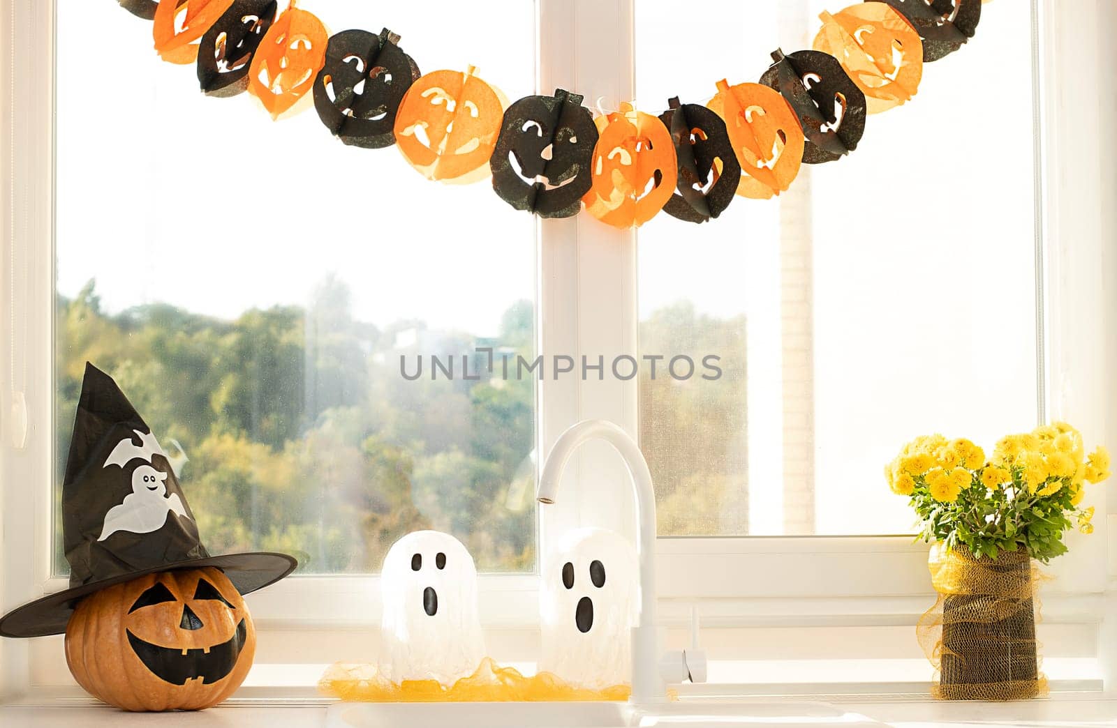 Halloween holiday concept. A pumpkin with a painted face, a white ghost and a bouquet of yellow chrysanthemum flowers in a black vase against the background of a window in home interior.