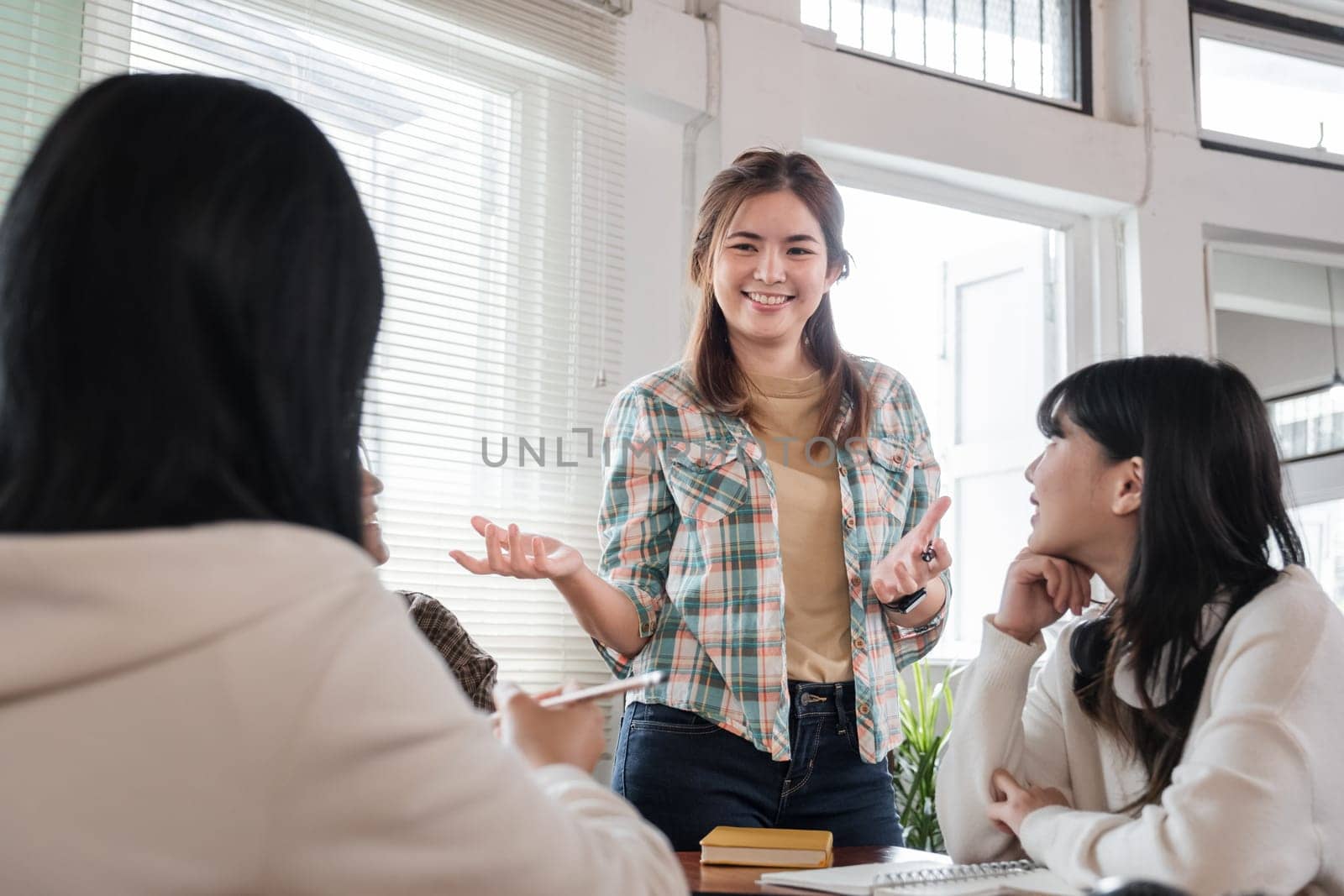 A cheerful and smart young Asian female is standing and sharing her ideas in a meeting with her team. University students, friendship, startups, teamwork by wichayada