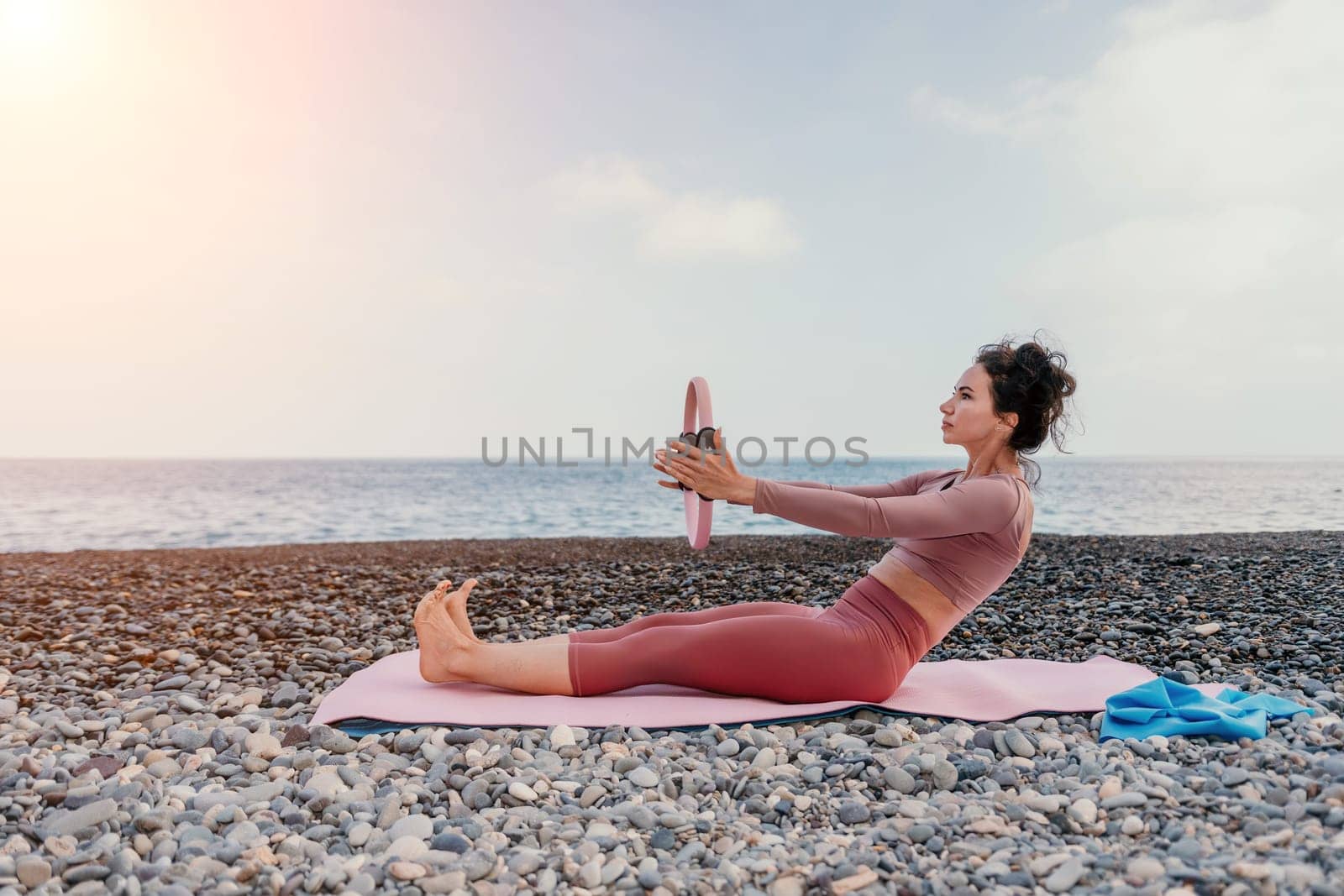 Middle aged well looking woman with black hair doing Pilates with the ring on the yoga mat near the sea on the pebble beach. Female fitness yoga concept. Healthy lifestyle, harmony and meditation.