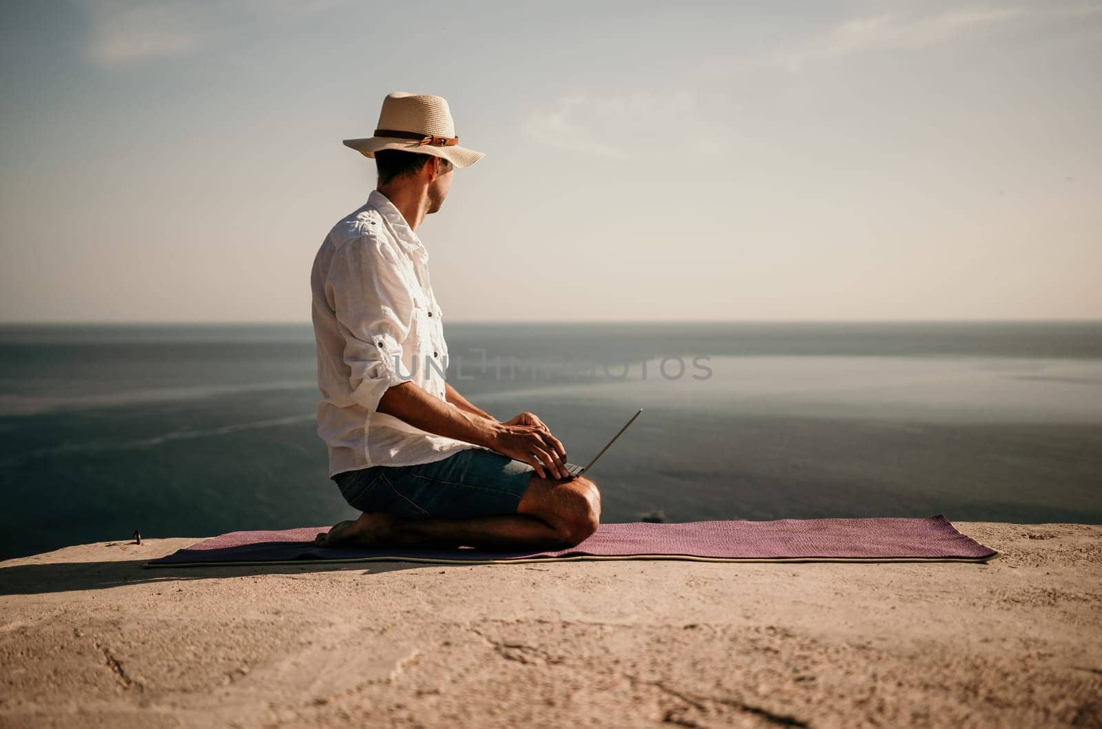 A digital nomad, a man in a hat, a businessman with a laptop does yoga on the rocks by the sea at sunset time, does a business operation online from a distance. Remote work on vacation. by panophotograph