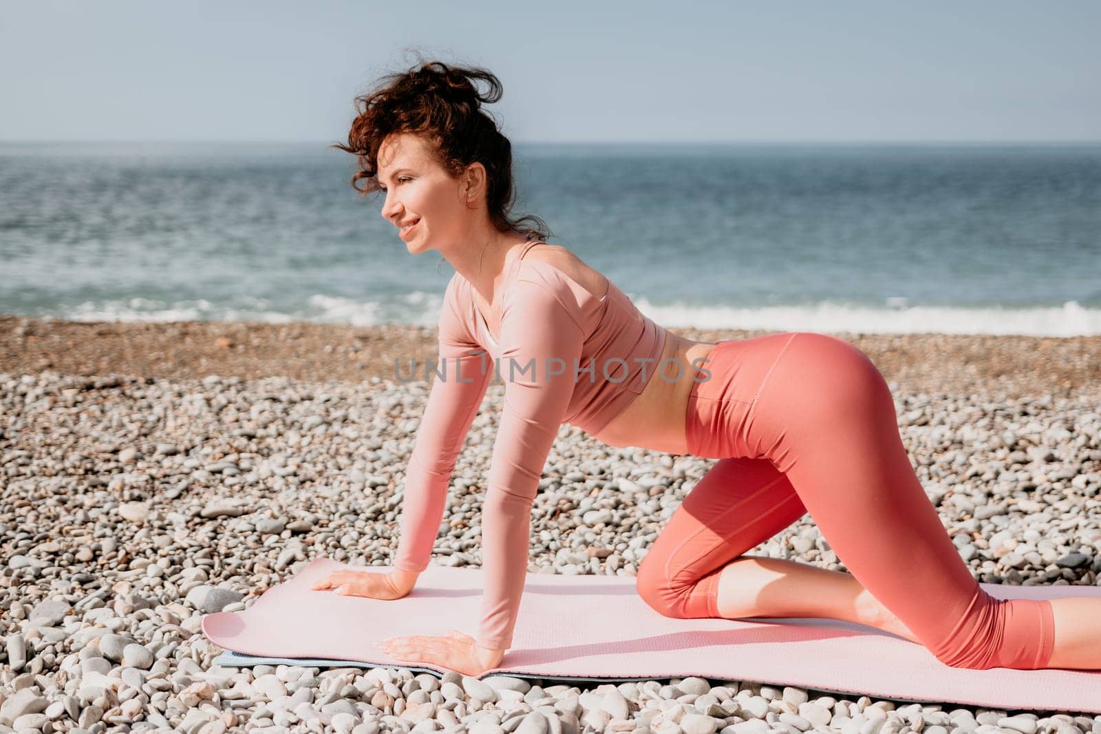 Middle aged well looking woman with black hair doing Pilates with the ring on the yoga mat near the sea on the pebble beach. Female fitness yoga concept. Healthy lifestyle, harmony and meditation.