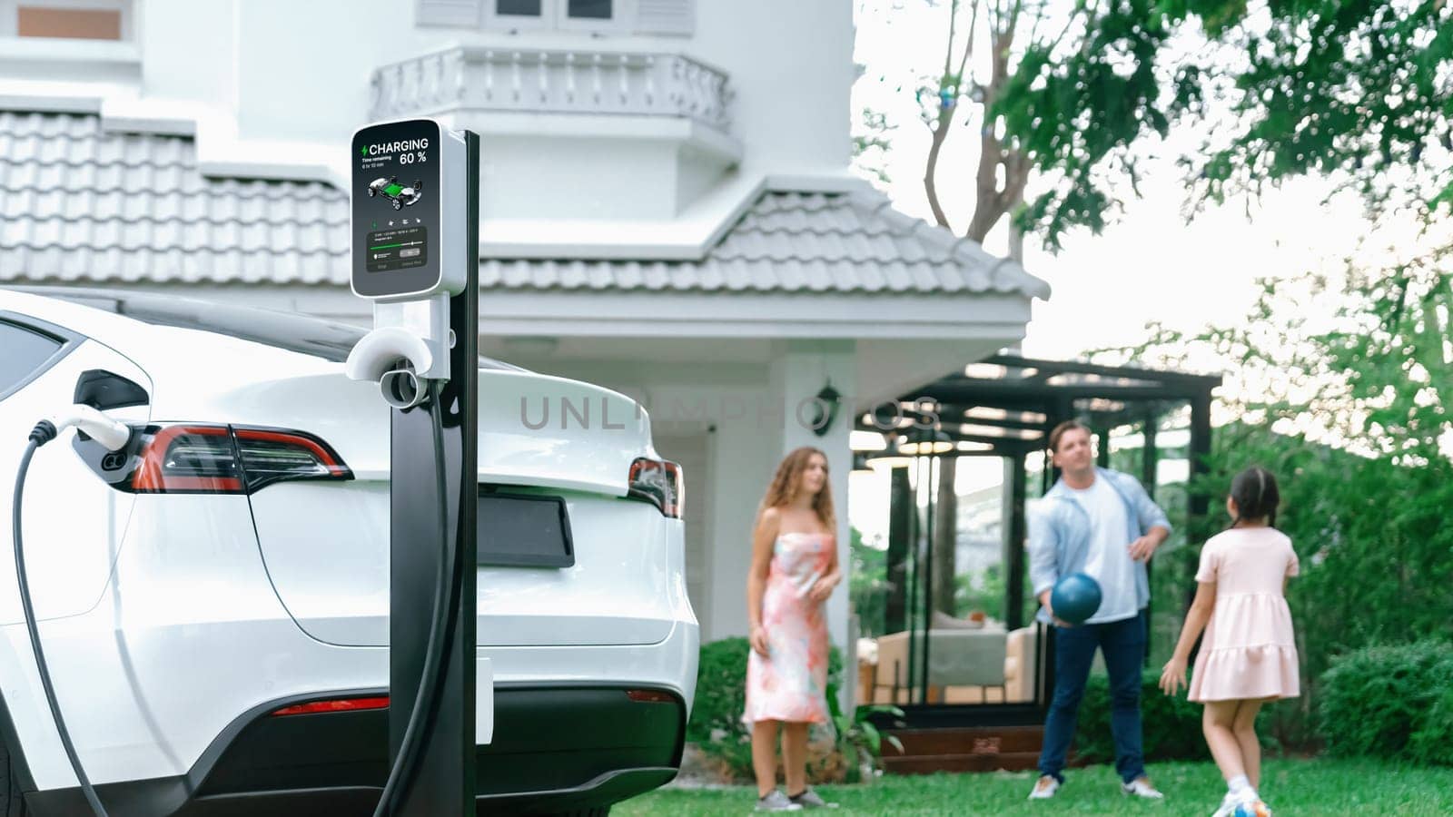 Focus electric vehicle recharge from home charging station on blur background of happy and playful family playing together. EV car using alternative and sustainable energy for better future.Synchronos