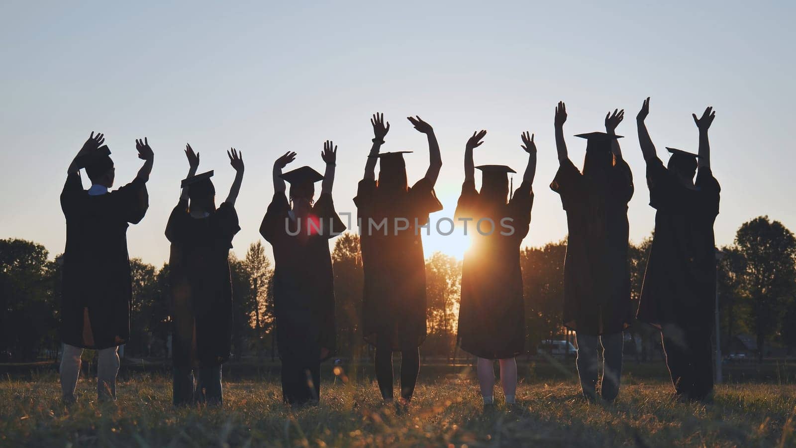College graduates in robes waving at sunset. by DovidPro