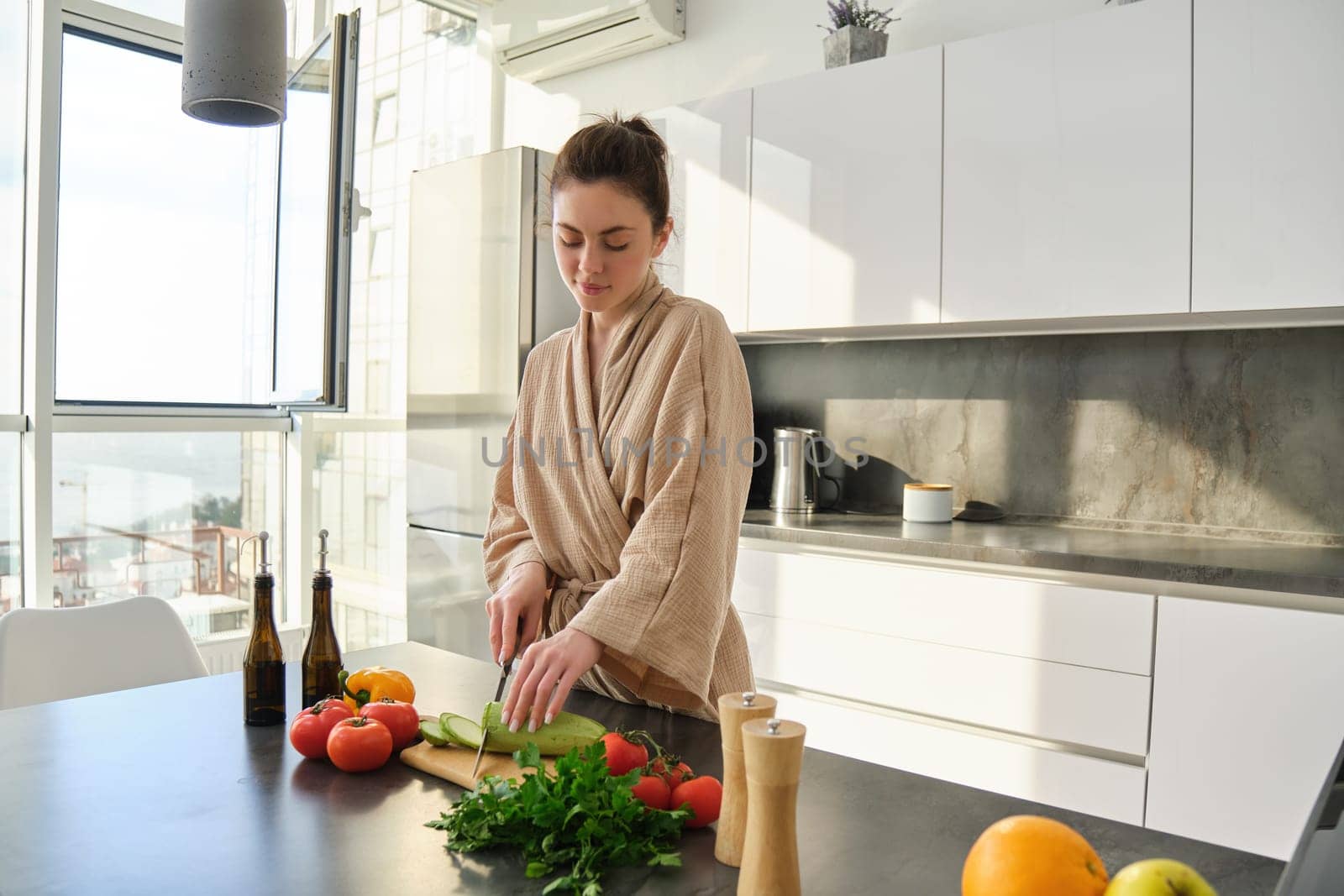 Portrait of woman cooking breakfast, chopping vegetables for salad, using board and knife, standing in the kitchen and wearing bathrobe.