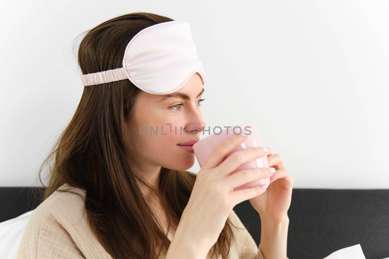 Portrait of beautiful woman relaxing in bed, waking up and having her morning cup of coffee in bedroom, looking happy by Benzoix