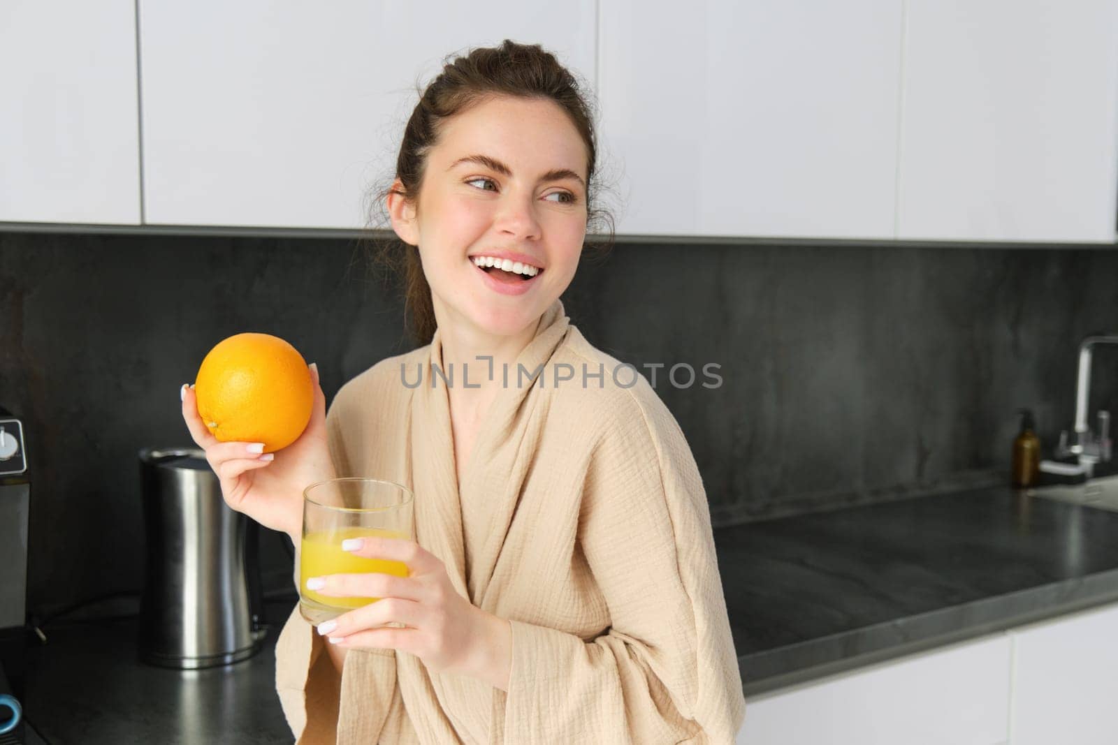 Portrait of good-looking brunette woman, drinking fresh orange juice in kitchen, holding fruit in hand, laughing and smiling, looking happy.