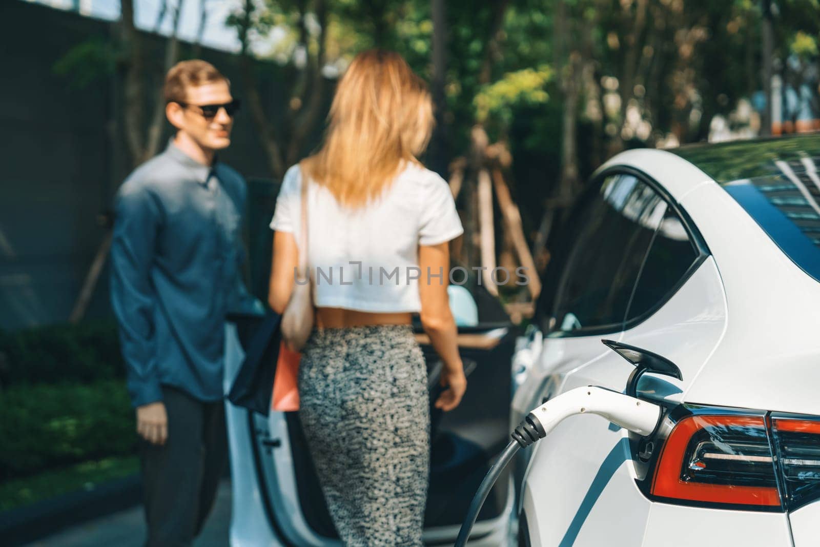 Young couple travel with EV electric car charging in green sustainable city outdoor garden in summer shows urban sustainability lifestyle by green clean rechargeable energy of electric vehicle innards