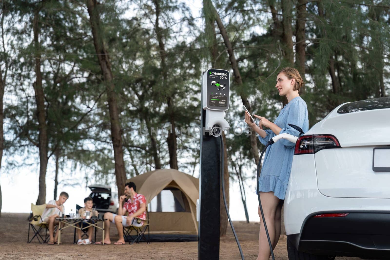 Outdoor adventure and family vacation camping at sea travel by eco friendly car. Woman or mother check car's battery with smartphone while charging EV car frin charging station in campsite. Perpetual